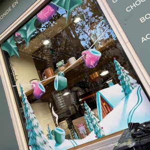 Idée déco Noël 2026 pour magasin : création artistique sur vitre avec forêt de sapins enneigés et village d’hiver féerique